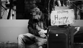 A homeless man sitting next to a sign and his luggage