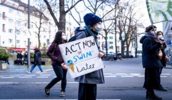 Woman in black jacket holding white printer paper photo saying "Act now or swim later."